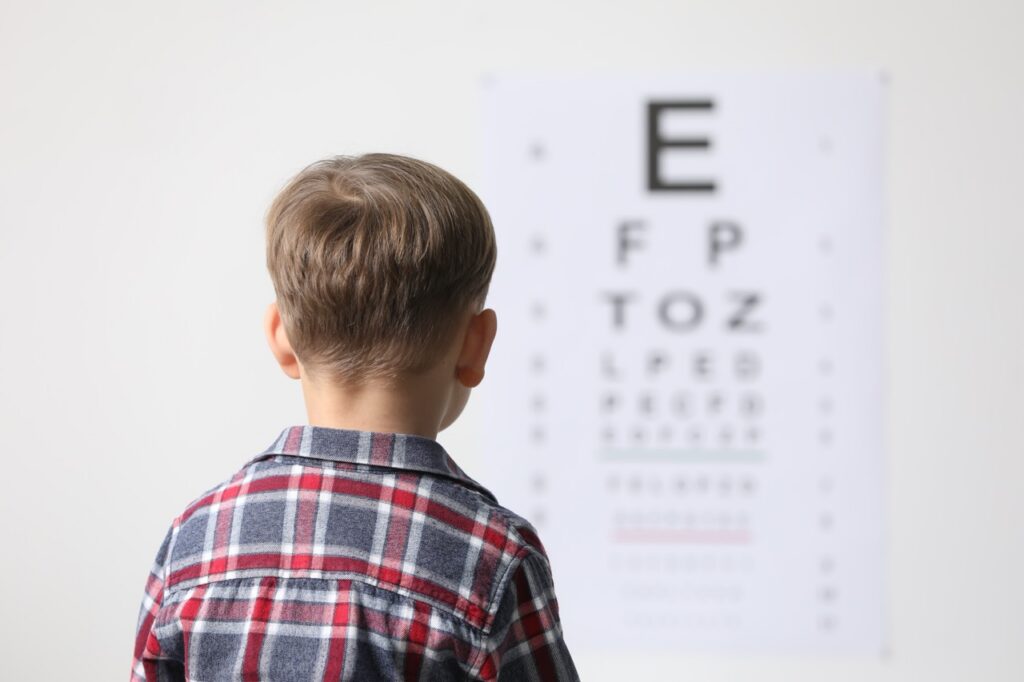 Young boy in a plaid shirt with a collar looking forward at a blurred out eye exam chart. How to know if your child has myopia