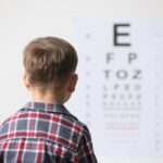 Young boy in a plaid shirt with a collar looking forward at a blurred out eye exam chart. How to know if your child has myopia