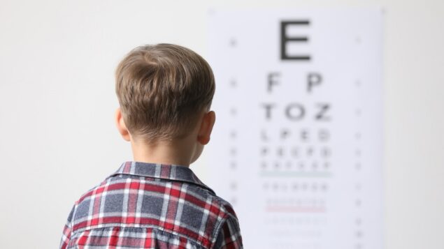 Young boy in a plaid shirt with a collar looking forward at a blurred out eye exam chart. How to know if your child has myopia