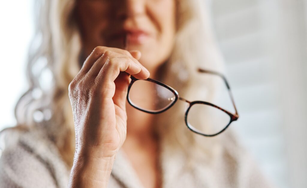 women with long light hair holding reading glasses. picture is cut off below her eyes and all is blurry except her hand and the glasses