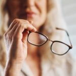 women with long light hair holding reading glasses. picture is cut off below her eyes and all is blurry except her hand and the glasses