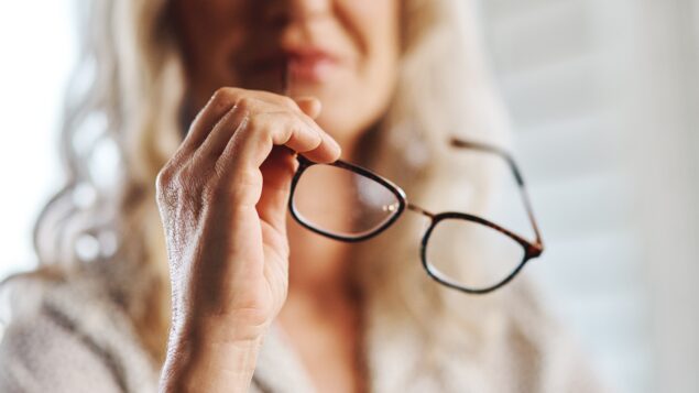 women with long light hair holding reading glasses. picture is cut off below her eyes and all is blurry except her hand and the glasses